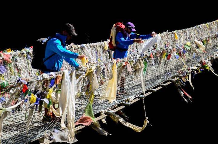 Dois caminhantes atam lenços khata brancos a uma ponte suspensa coberta de bandeiras de oração.
