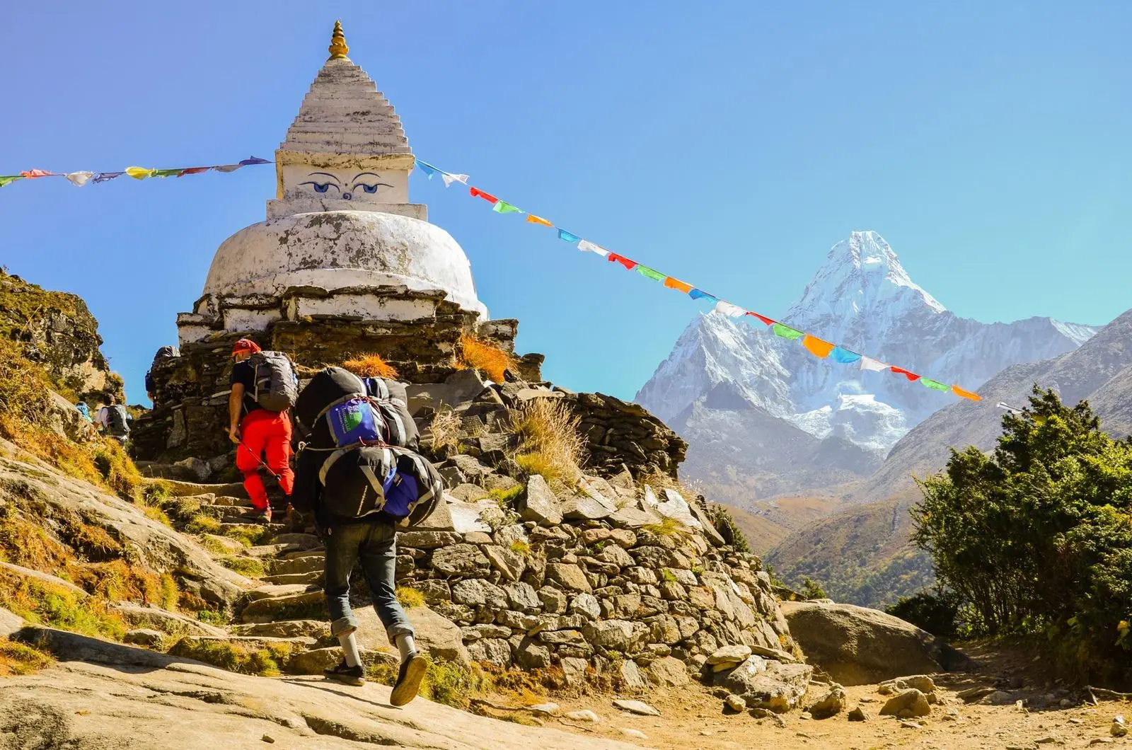 Um carregador e um caminhante passam por uma stupa com os olhos de Buda; à direita ergue-se o Ama Dablam.